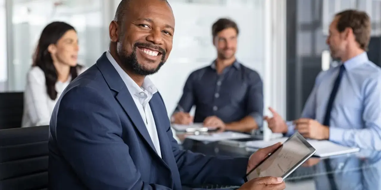 The executive board chair smiles, holding a tablet in a meeting with colleagues.